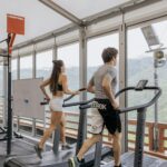 Man and woman running on treadmills in a bright gym with scenic views.